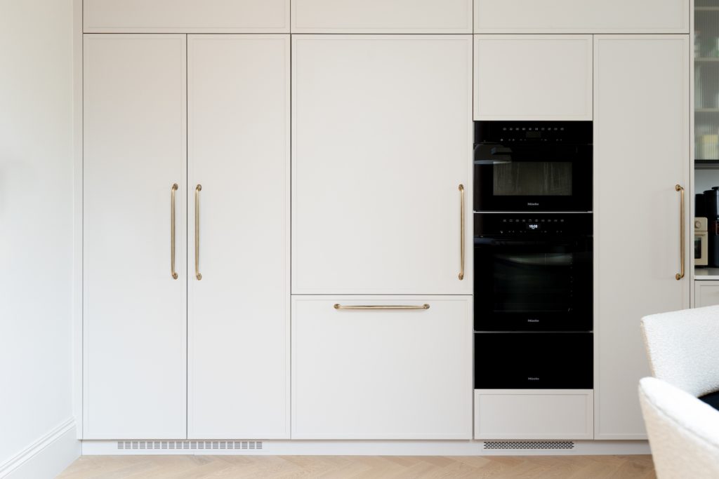 Full-height pale bespoke kitchen cabinetry with integrated ovens and refined metallic handles in the Oakhill project.